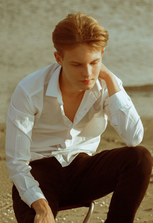 Free Contemplative male sitting on beach in white shirt and black pants. Summer vibe. Stock Photo