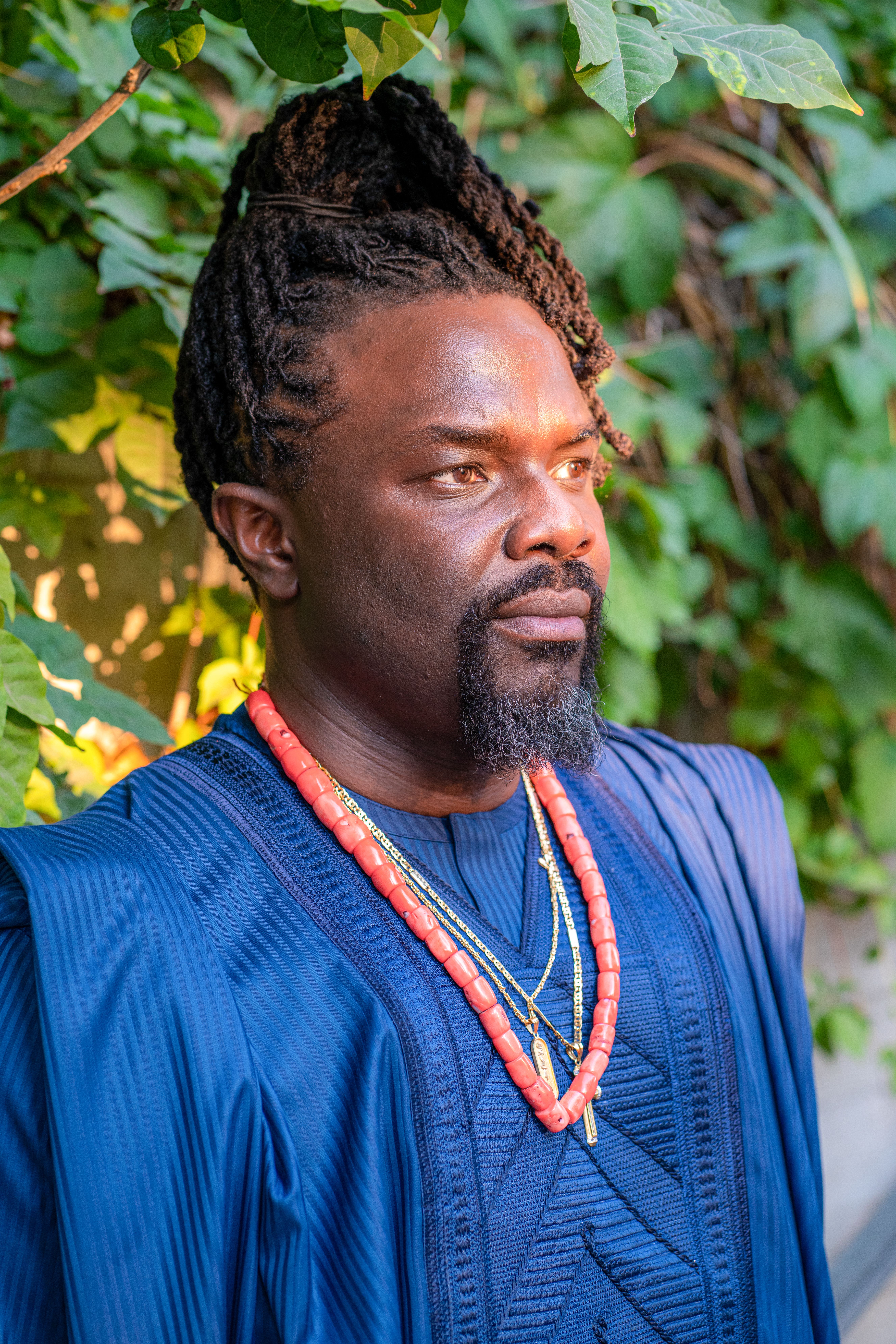 A portrait of a man in blue traditional clothing with braided hair and necklaces, set outdoors.