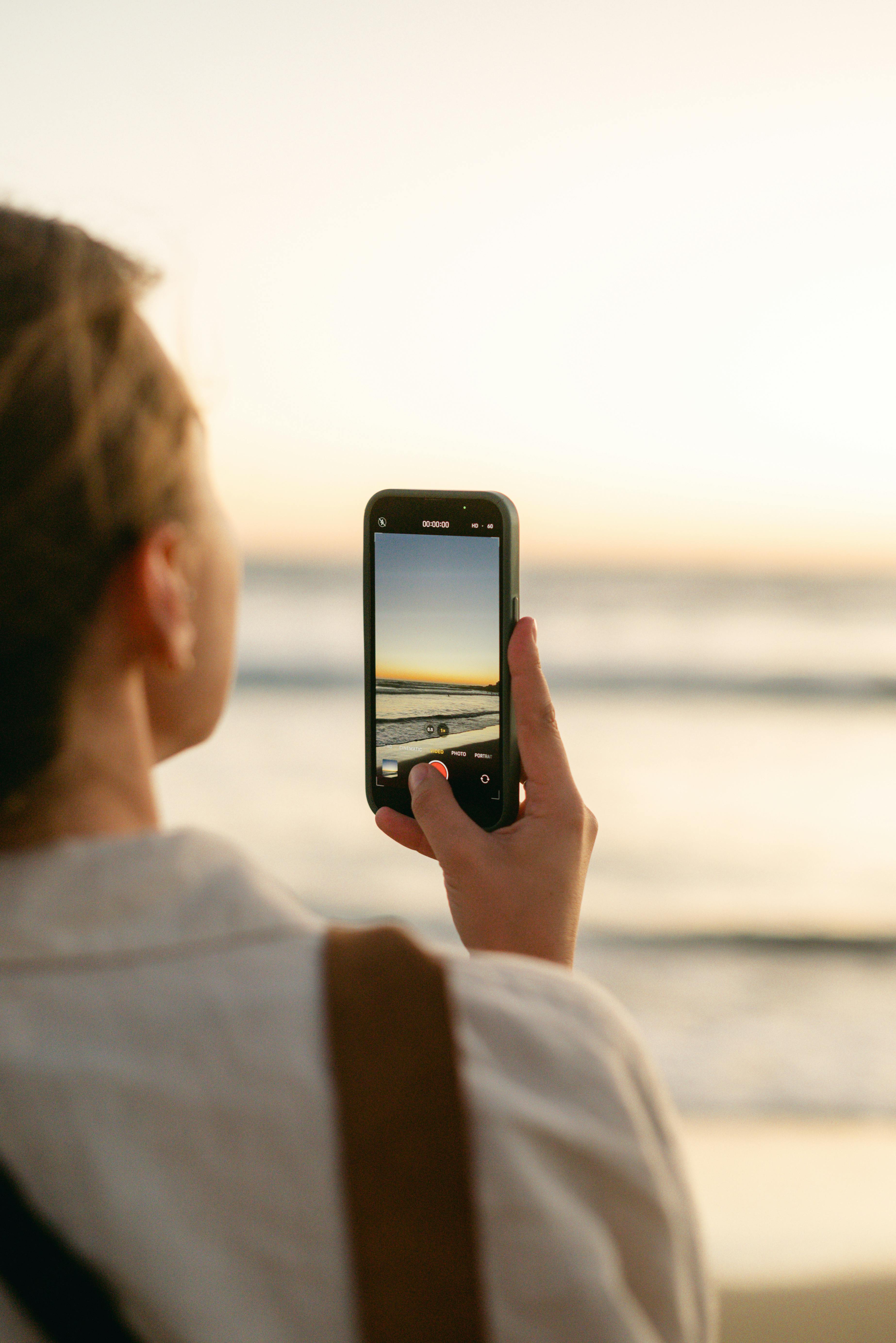 A woman photographs a peaceful sunset on her smartphone at the beach, embracing the moment.