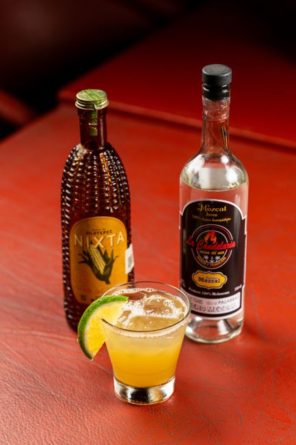 A bottle of mezcal next to a small clay cup, lime wedges, and dried worm salt on a wooden table.