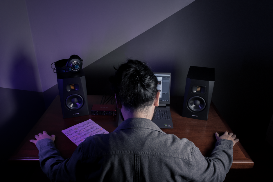 Home recording setup viewed from behind, showing studio monitors, an audio interface, headphones, and a laptop on a wooden desk.