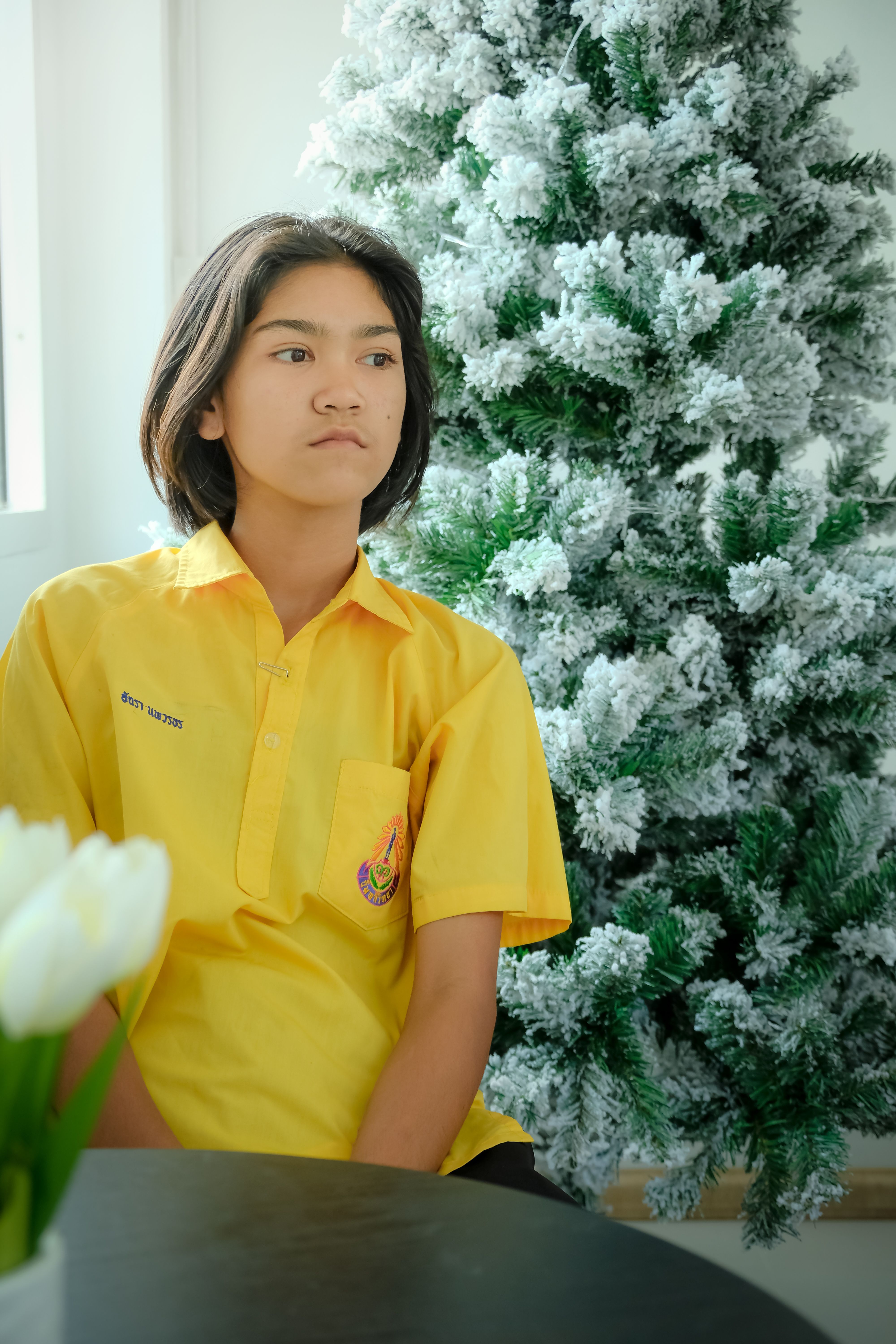 Young girl sits beside a frosted Christmas tree wearing a yellow shirt, in a bright indoor setting.