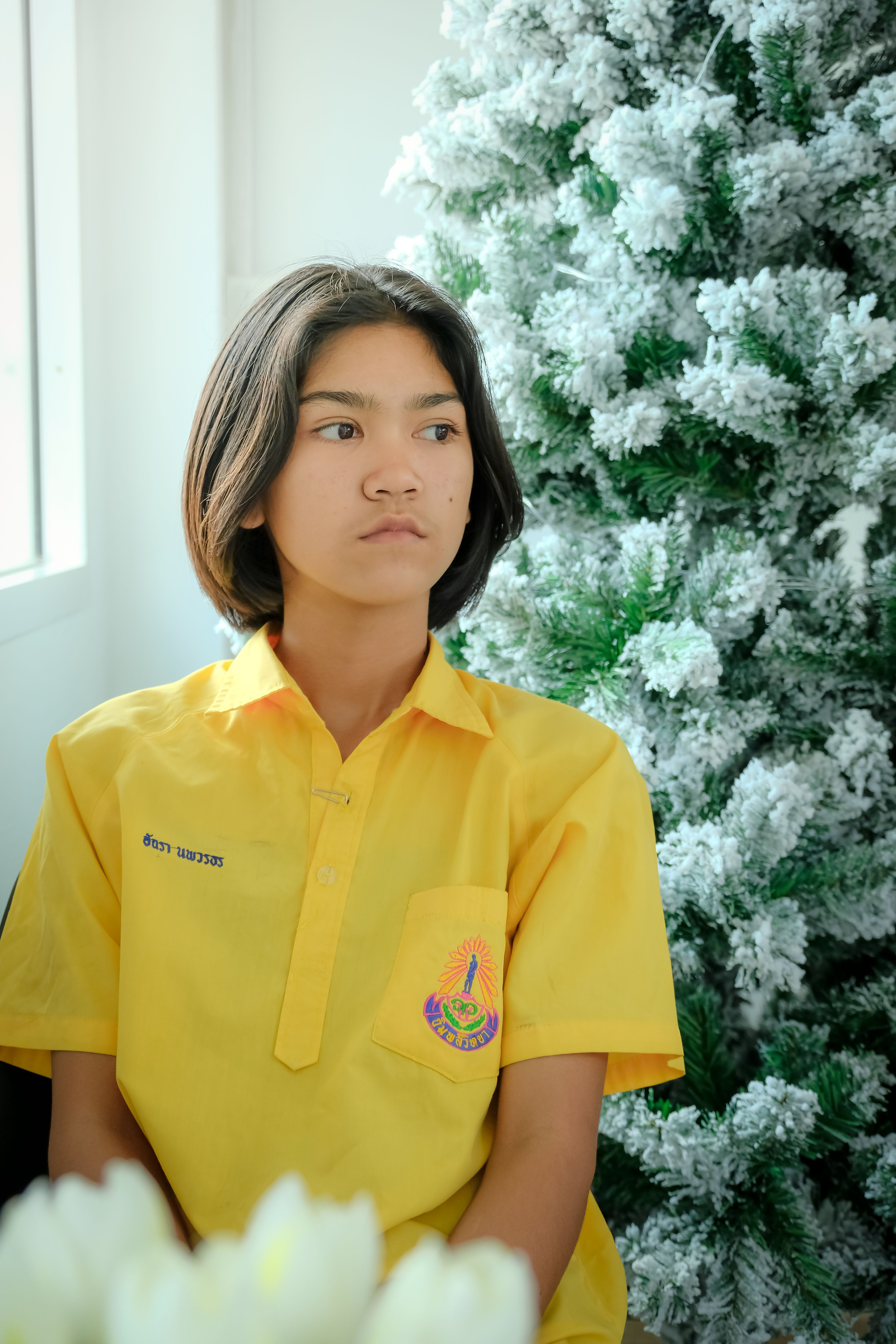 A contemplative young girl in a yellow shirt sits by a frosted Christmas tree indoors.