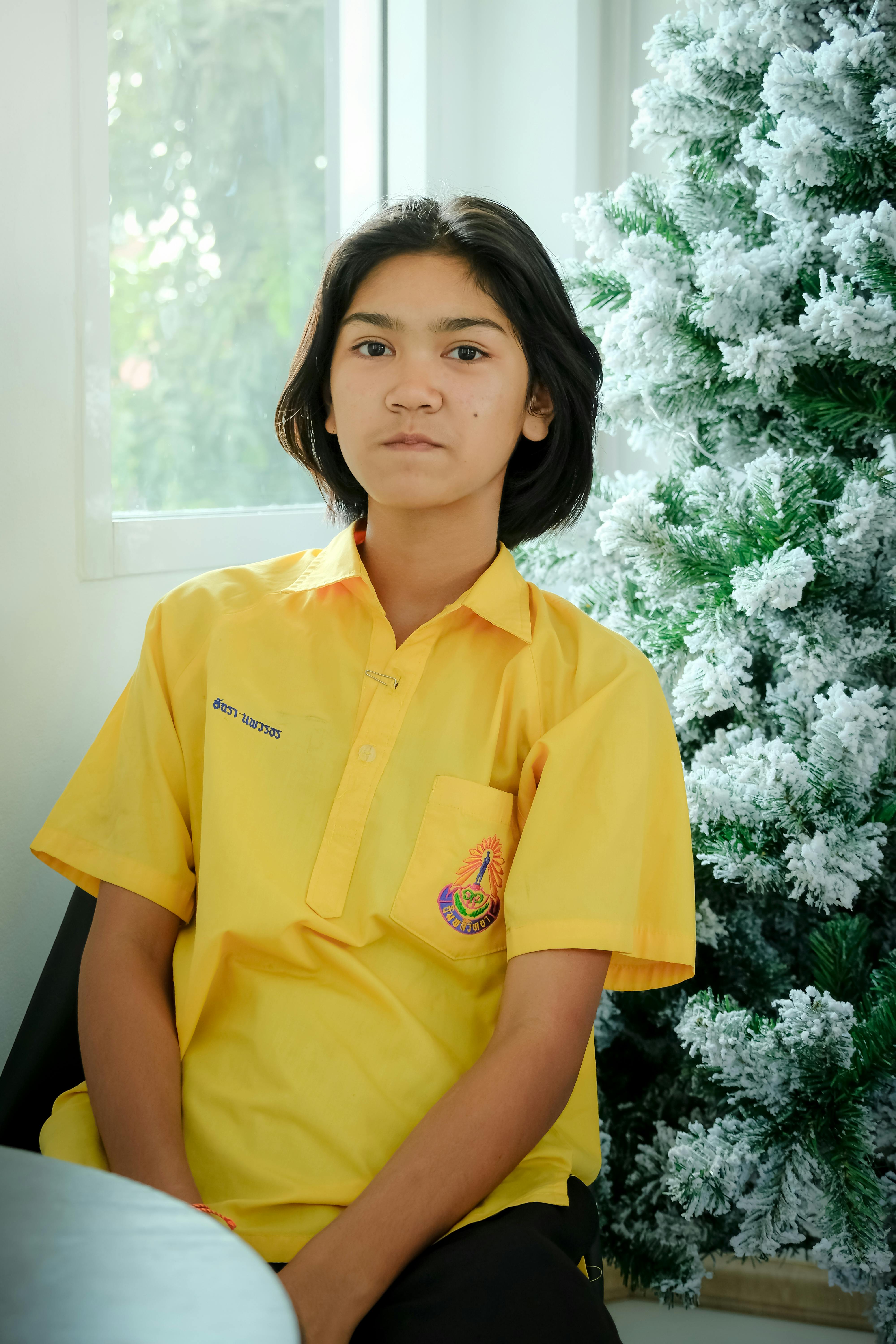 Portrait of an Asian teenage girl in yellow shirt sitting by a decorated Christmas tree.