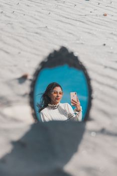 Woman taking a selfie reflected in a mirror on a sunny beach day.