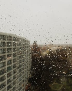 Captured raindrops on windowpane with urban cityscape view, creating a moody atmosphere.