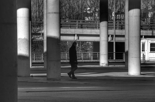Free Black and white photo of a man walking alone in a city under concrete pillars. Stock Photo