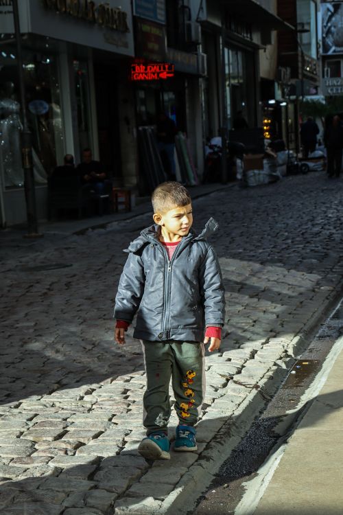 Free Young boy in jacket stands on a cobbled street in a vibrant urban area, illuminated by sunlight. Stock Photo