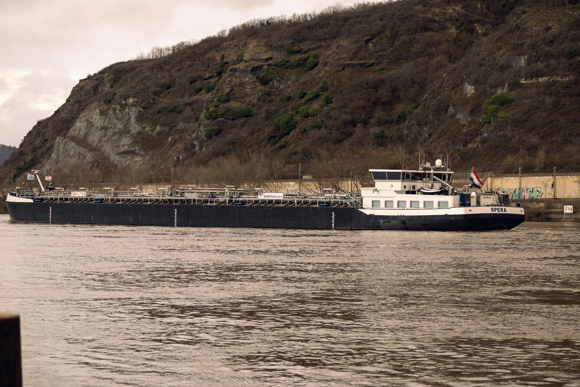 A large cargo ship navigates a calm river with a rocky hill backdrop, exemplifying industrial transport.