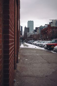 A snowy city sidewalk lined with parked cars and buildings on a cloudy winter day.