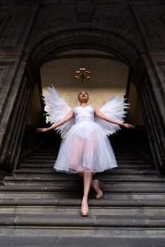 A graceful ballerina in a white dress with angel wings poses on grand stairs.