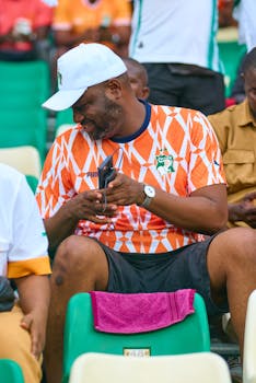 Black man in Ivory Coast soccer jersey smiling at stadium, holding phone.