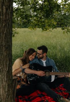 A loving couple enjoys a summer day in the meadow, playing guitar under a tree.