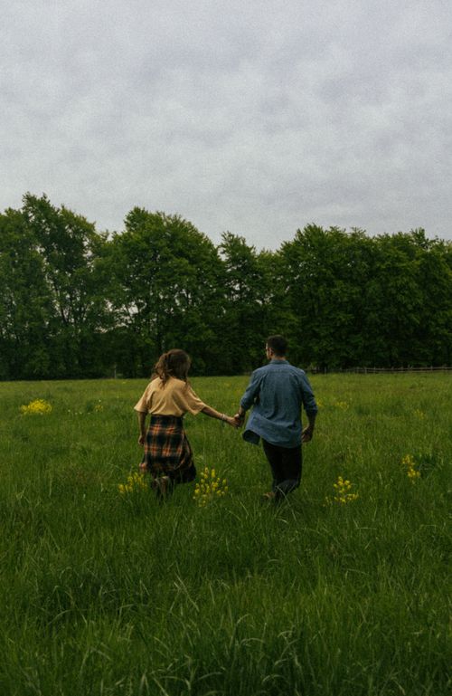 Free A couple holding hands and walking through a lush green meadow, surrounded by trees. Stock Photo