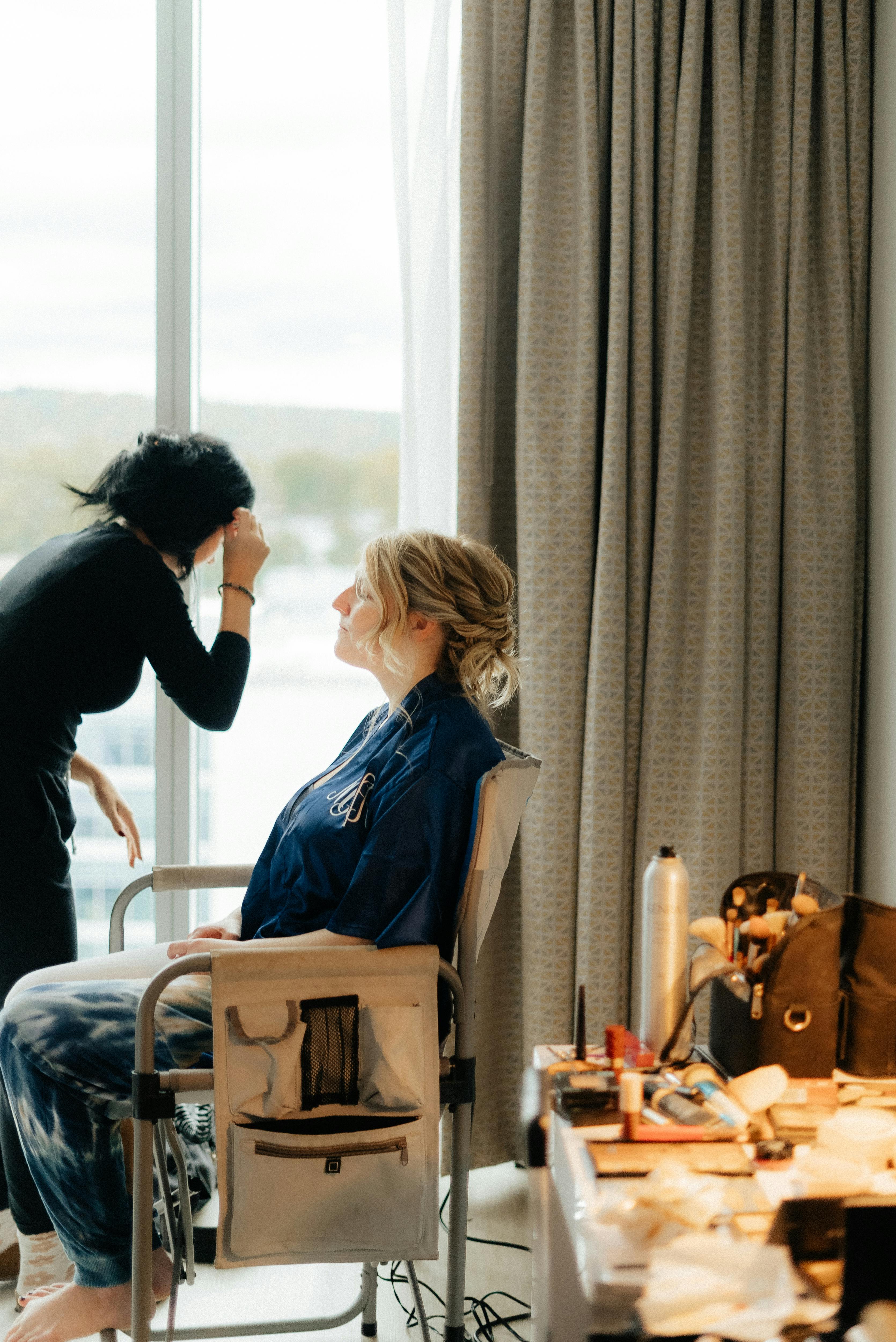 Makeup artist working on client near window, creating an elegant look indoors.