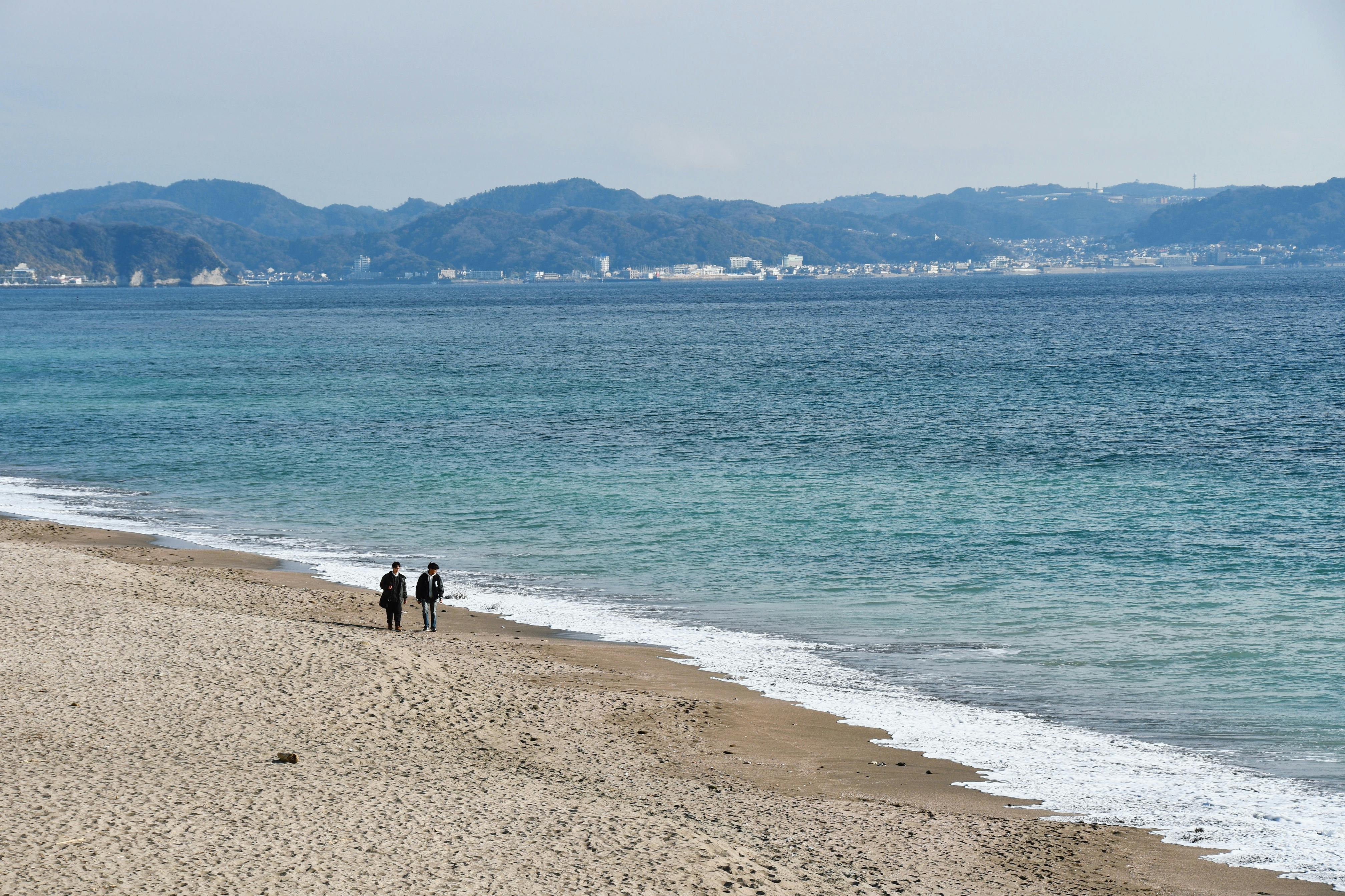 Two people walk along a serene beach with distant mountains across the ocean.