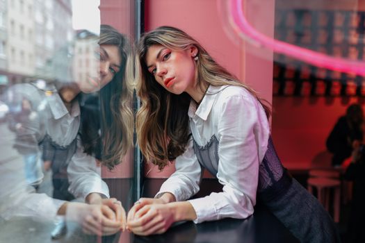 Fashionable woman leaning against a reflective window in a modern urban setting with neon lighting.