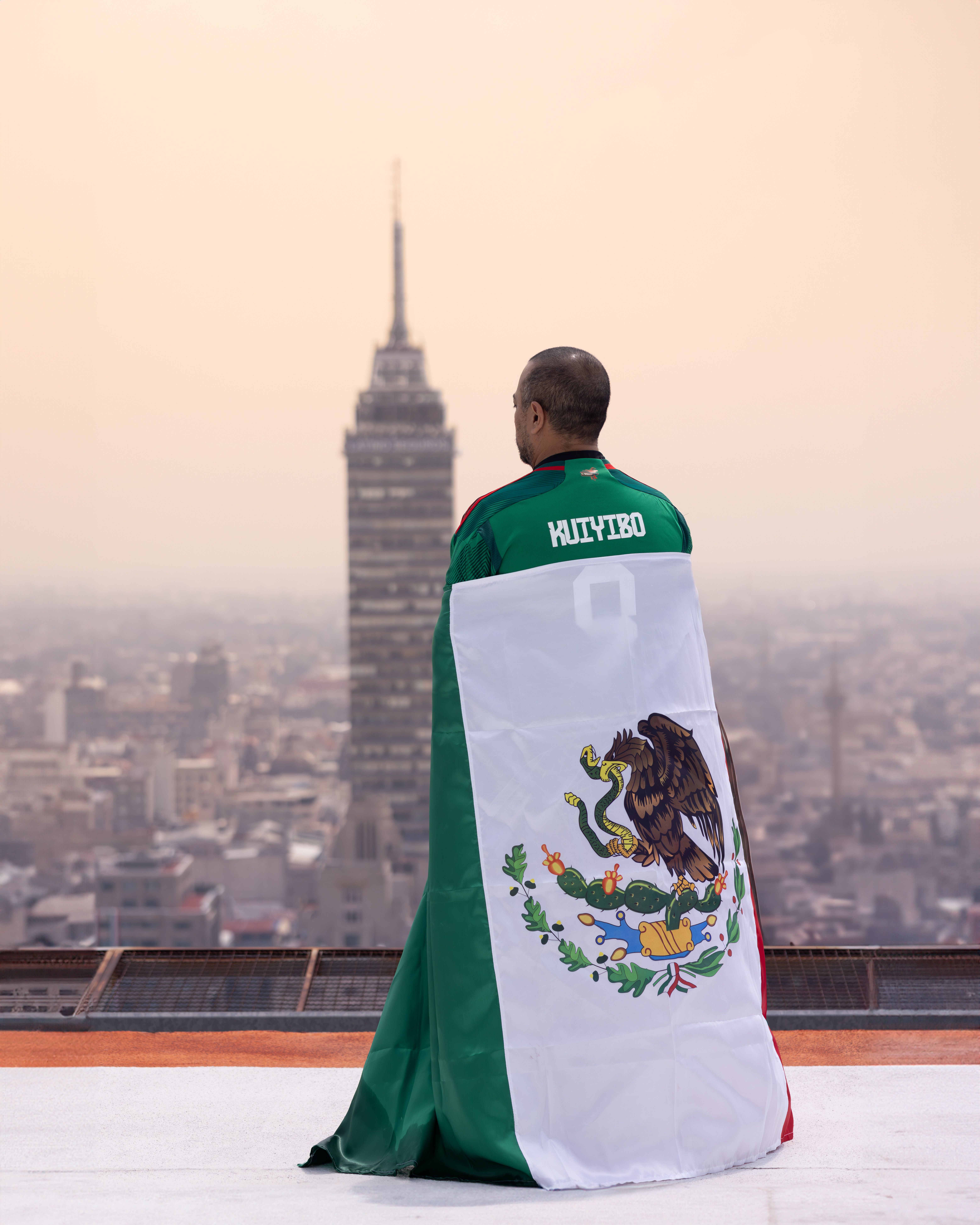 A man stands wrapped in a Mexican flag gazing at the skyline of Mexico City, featuring the Torre Latinoamericana tower.