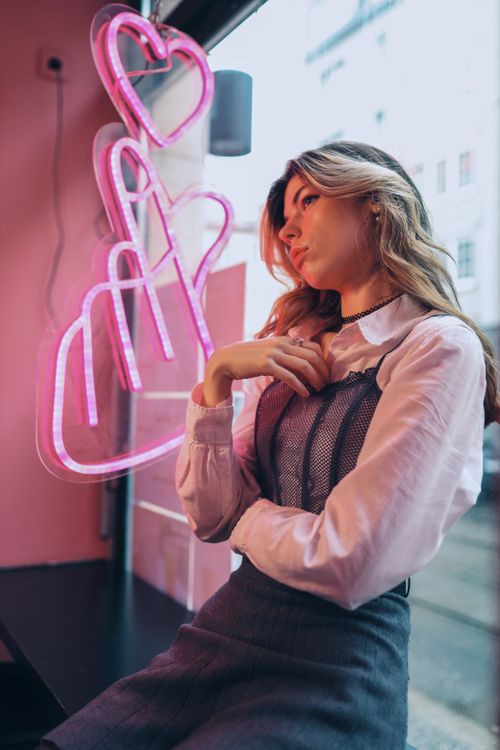Free Trendy portrait of a young woman sitting by neon lights in a Berlin cafe, with a stylish outfit and thoughtful expression. Stock Photo