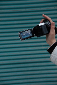 Close-up of a person's hand holding a Handycam with an LCD screen outdoors against a striped background.