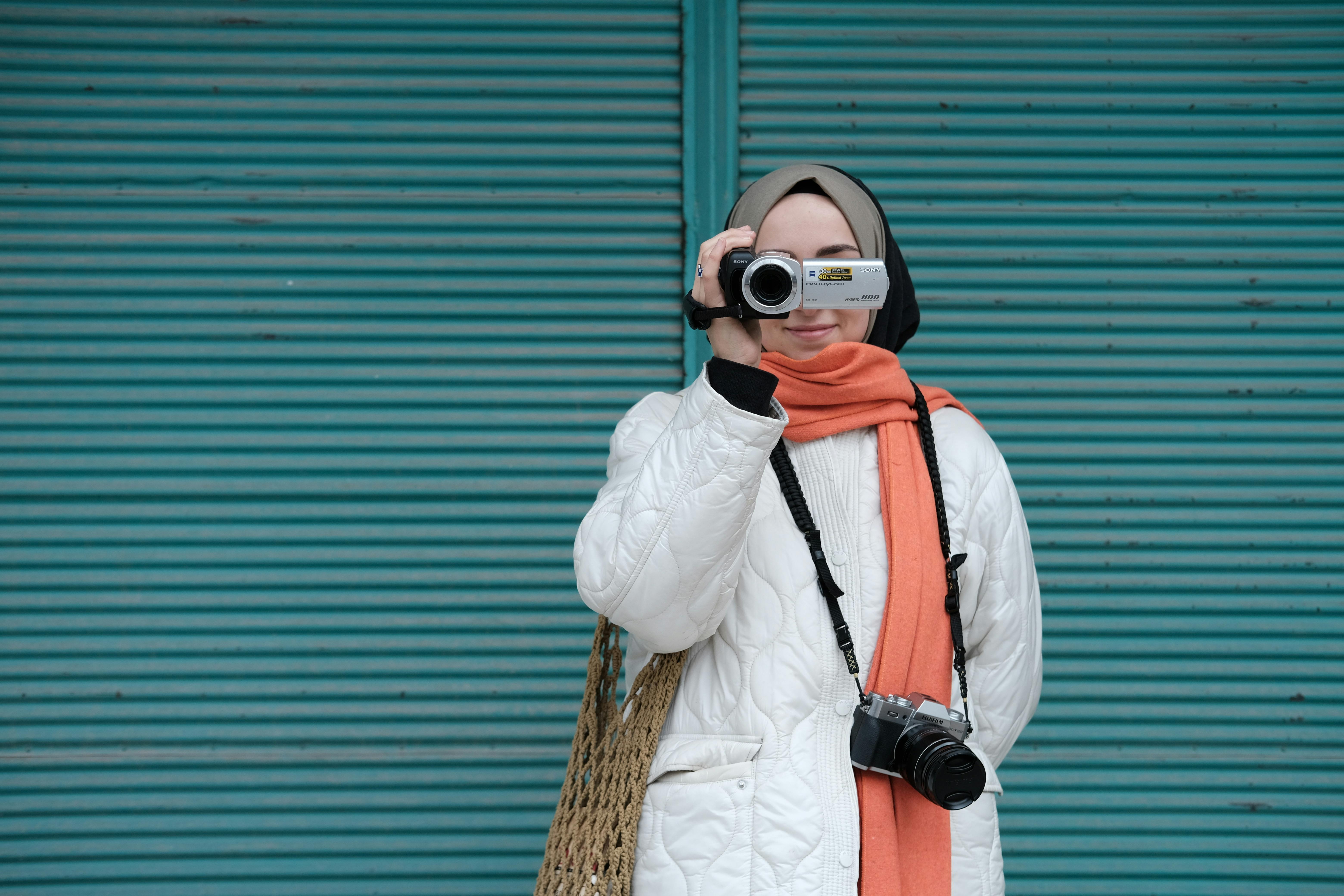 A young woman in a hijab records video with a handycam against a turquoise background.