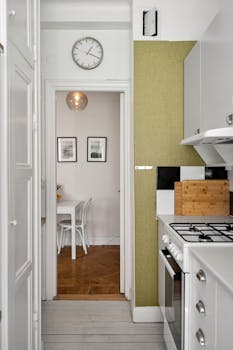Contemporary narrow kitchen leading to a cozy dining area. Clean design with white and green accents.