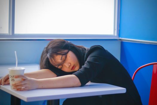 A woman sleeping on a café table, holding a disposable coffee cup, looking tired.