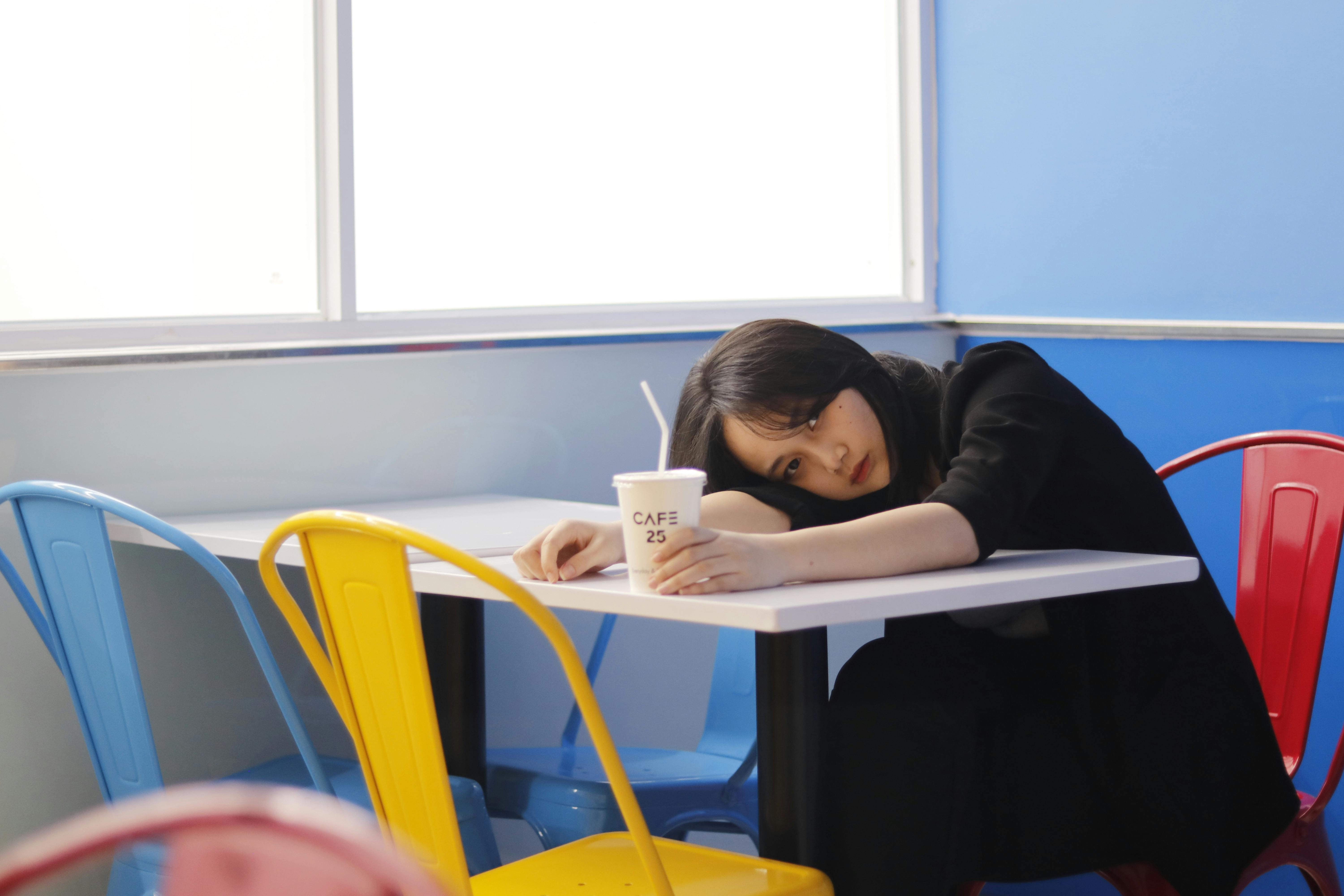 A young woman in a café, resting her head on a table with a drink in hand, surrounded by colorful chairs.