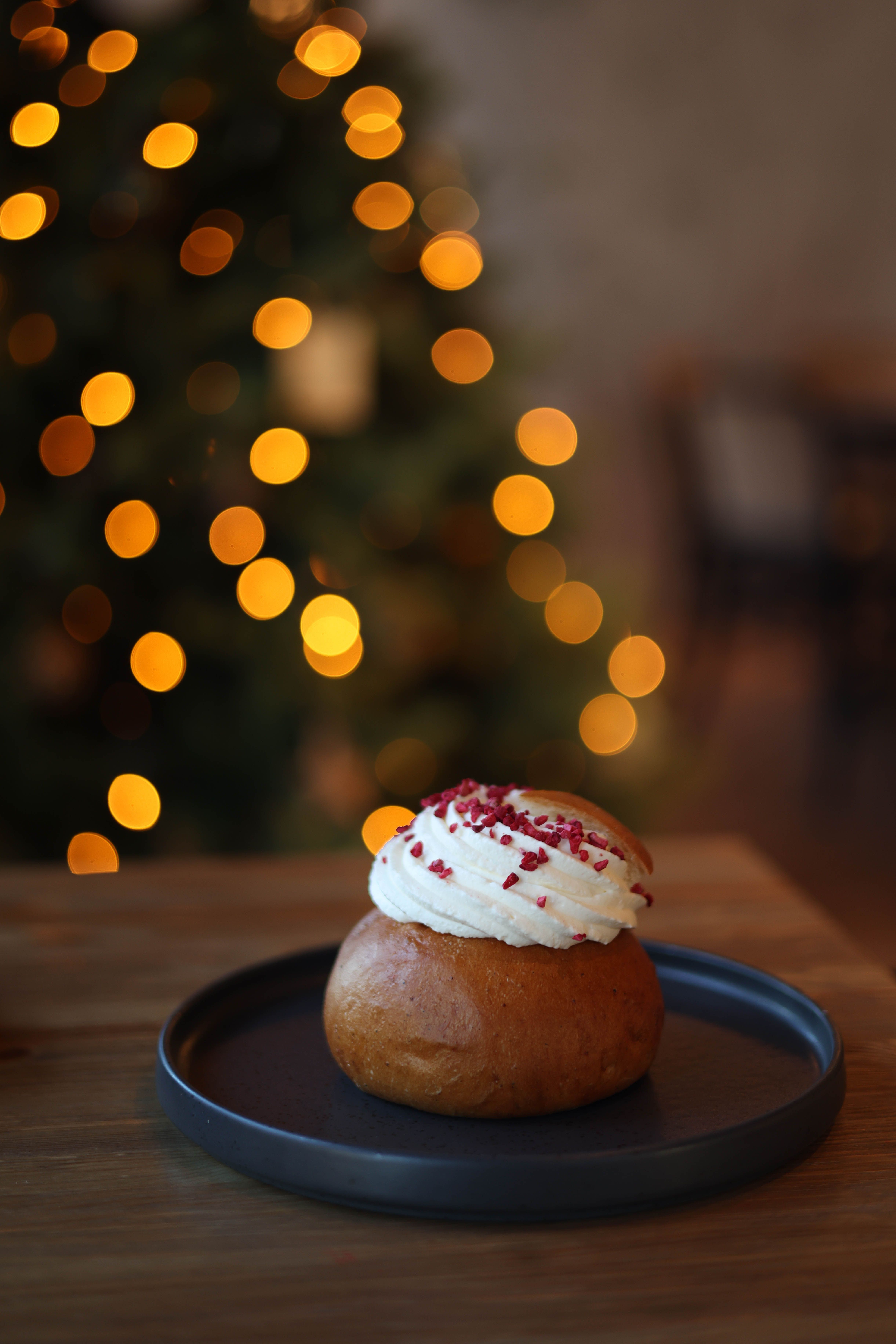A traditional Swedish semla bun topped with whipped cream, captured next to a Christmas tree with bokeh lights.