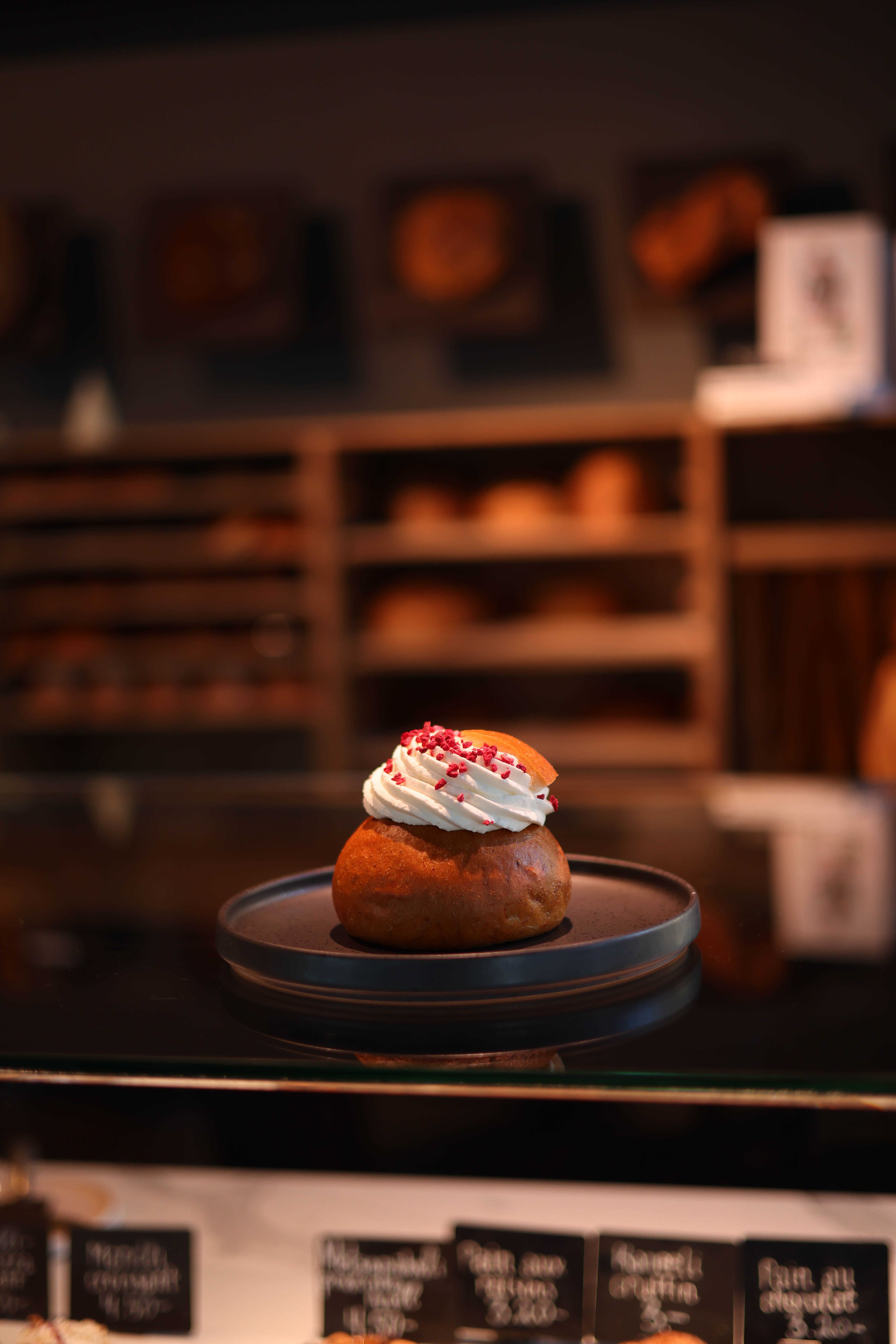 A delectable cream pastry topped with berries on a black plate, showcased in a bakery setting.