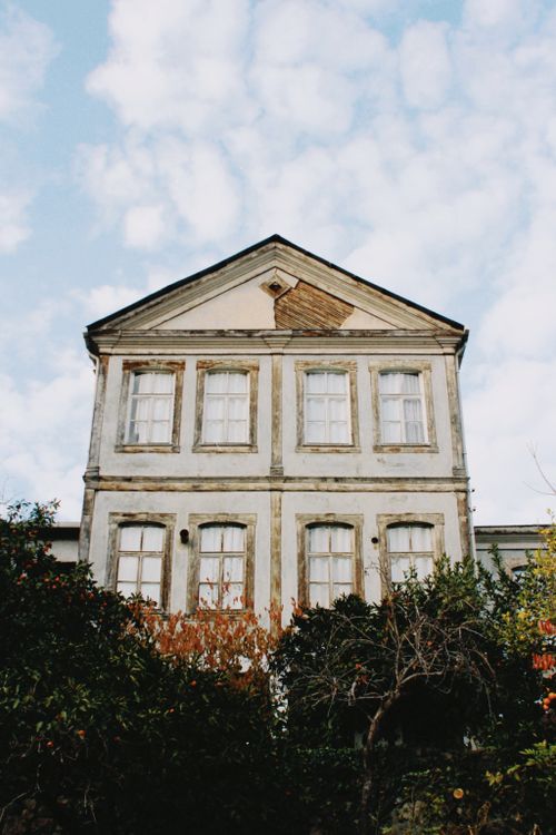 Free Old rural house facade with decaying textures, framed by trees and a blue sky with clouds. Stock Photo
