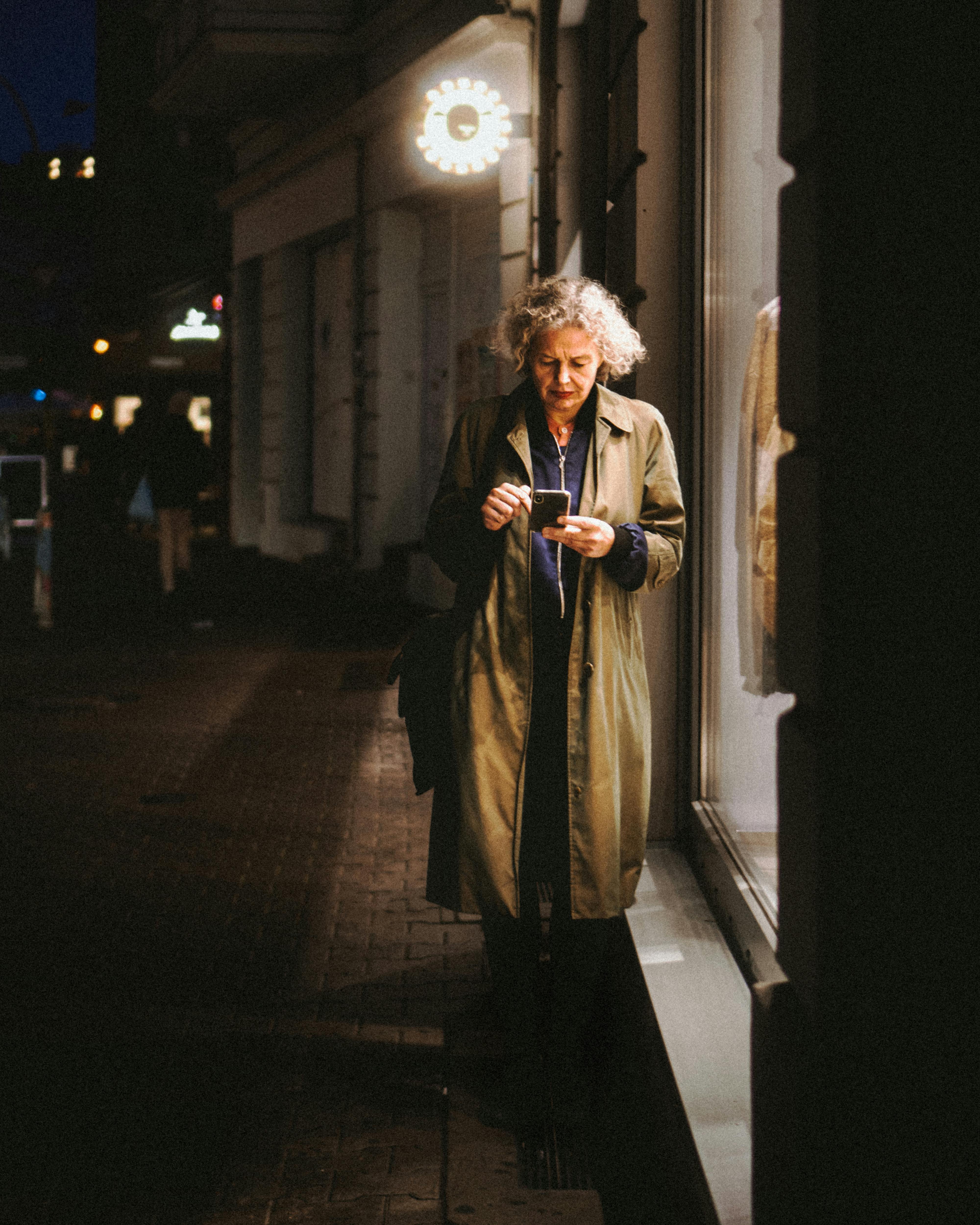 An elderly woman using her phone on a Berlin street at night, illuminated by store lights.