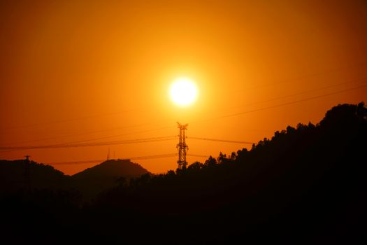 A striking silhouette of a transmission tower against a vibrant sunset sky with hills in the background.