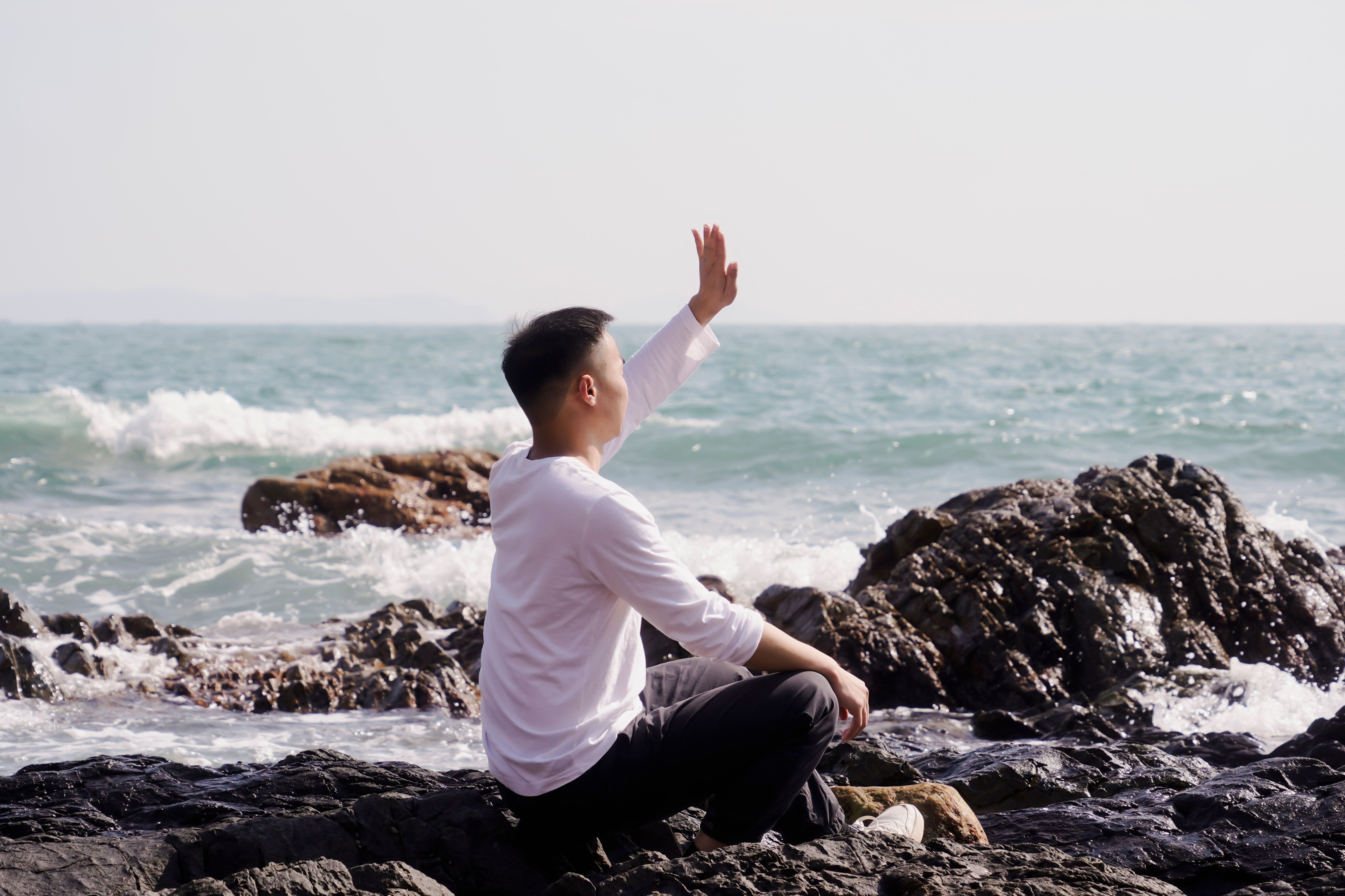 A man sitting on a rocky shore, waving towards the ocean under bright daylight.