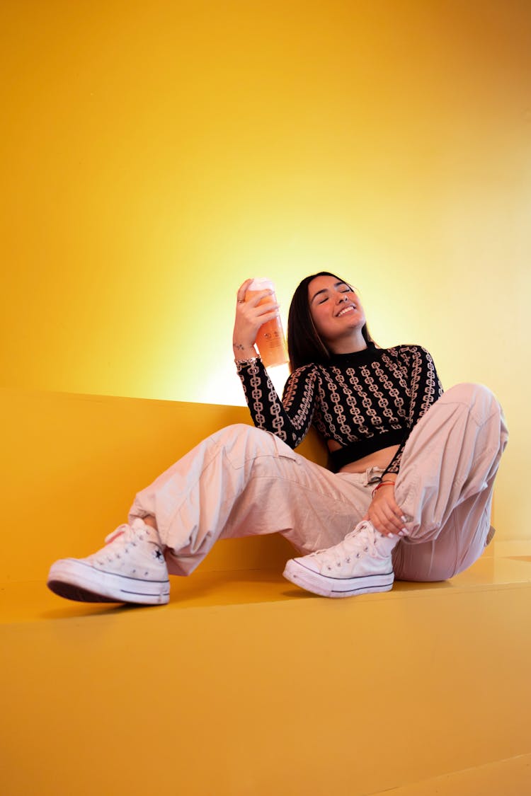 Happy Woman In Beige Pants Sitting In Studio With Orange Drink In Hand