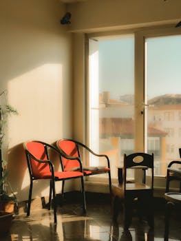 Cozy interior space with sunlit red chairs and large windows, overlooking buildings.