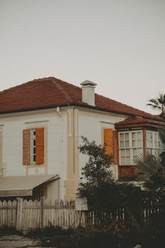 A beautifully preserved vintage house with a red tiled roof, surrounded by greenery and a wooden fence.