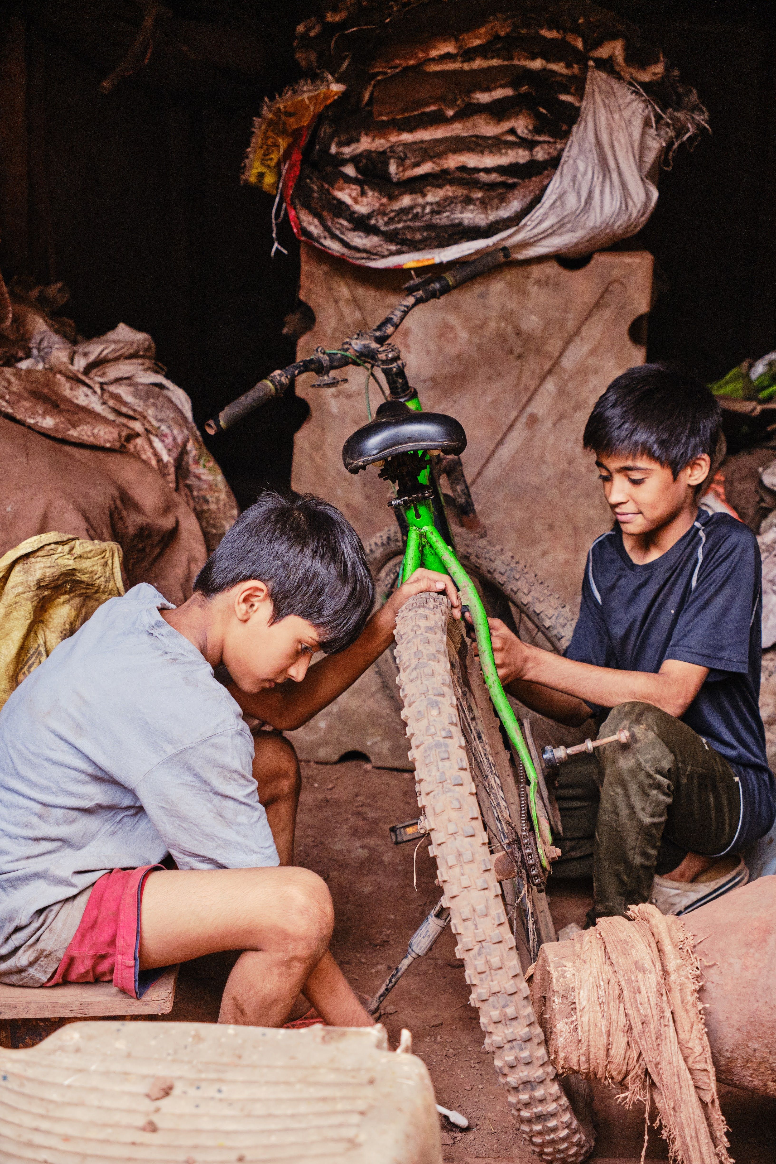 Two boys focus on fixing a bicycle in a makeshift outdoor workshop, surrounded by rustic materials.