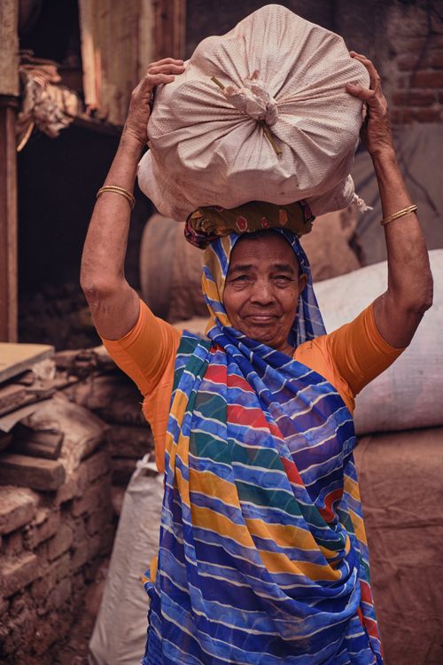 Free Portrait of an elderly woman in a colorful saree carrying a sack on her head, set in an outdoor rustic setting. Stock Photo