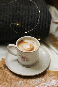 A floral coffee cup with creamy art on a saucer, surrounded by cozy decor and fairy lights.