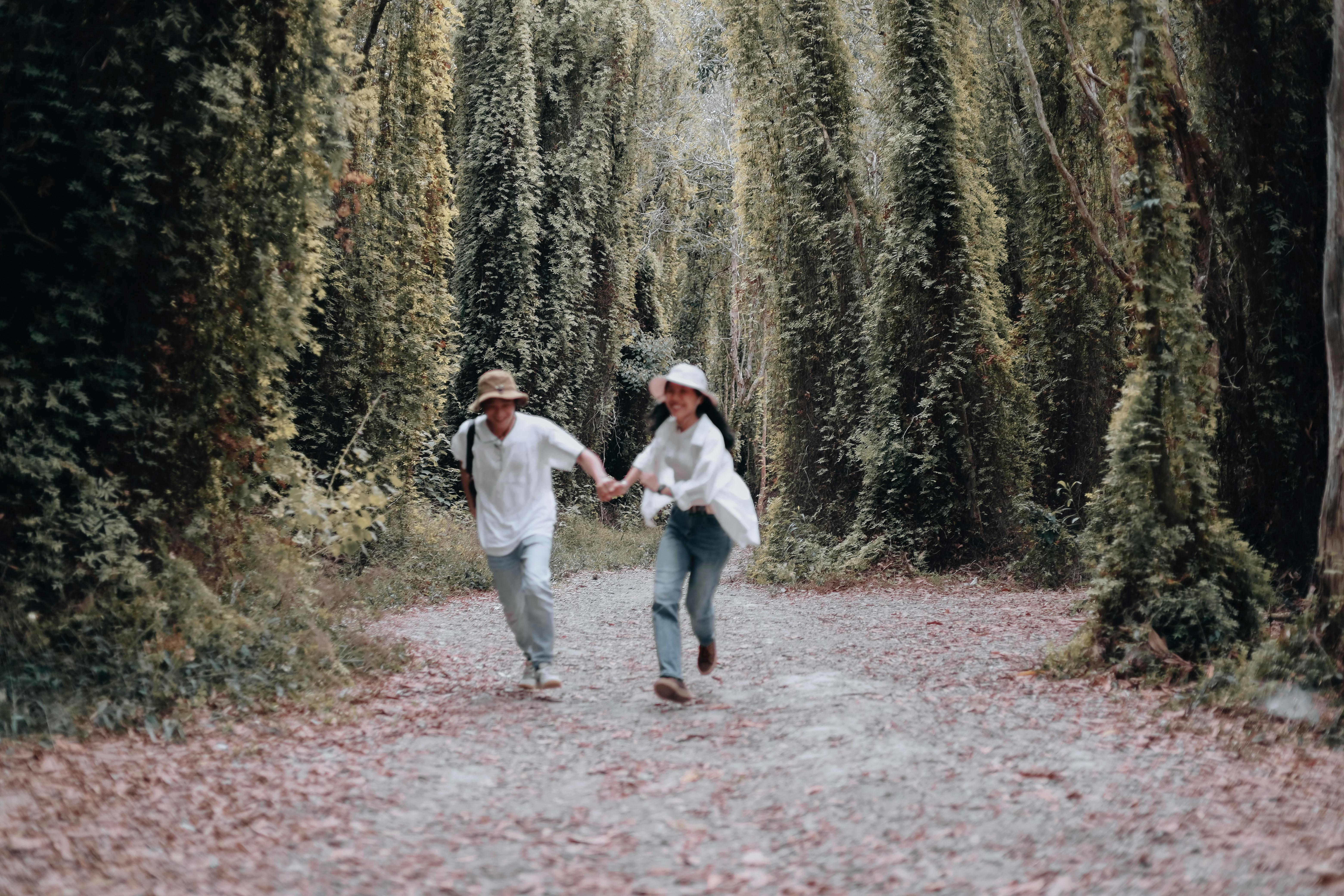 A couple runs joyfully through a lush forest, surrounded by tall trees.