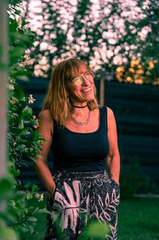 Smiling woman with glasses enjoys a summer sunset in a garden, wearing a floral skirt.