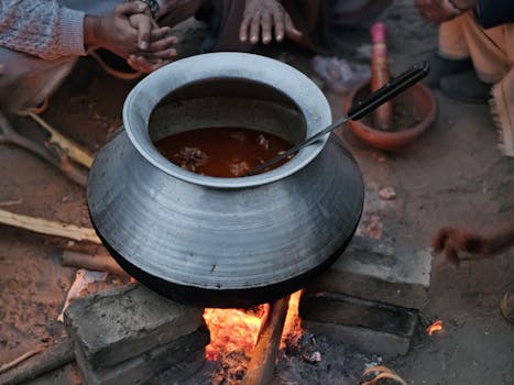 A large silver pot simmering stew over a bonfire, surrounded by people in a rural setting.