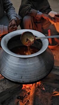 A traditional outdoor pot cooking over an open fire with a kitchen spoon and people gathered around.