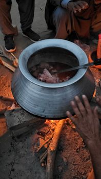 A large silver pot cooking over an outdoor bonfire, surrounded by people.