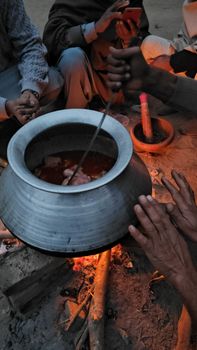 A group of people cooking outdoors in a large silver pot over a bonfire, sharing a communal meal.