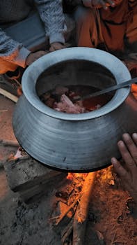 A group of people cooking meat in a silver pot over a bonfire at dusk.