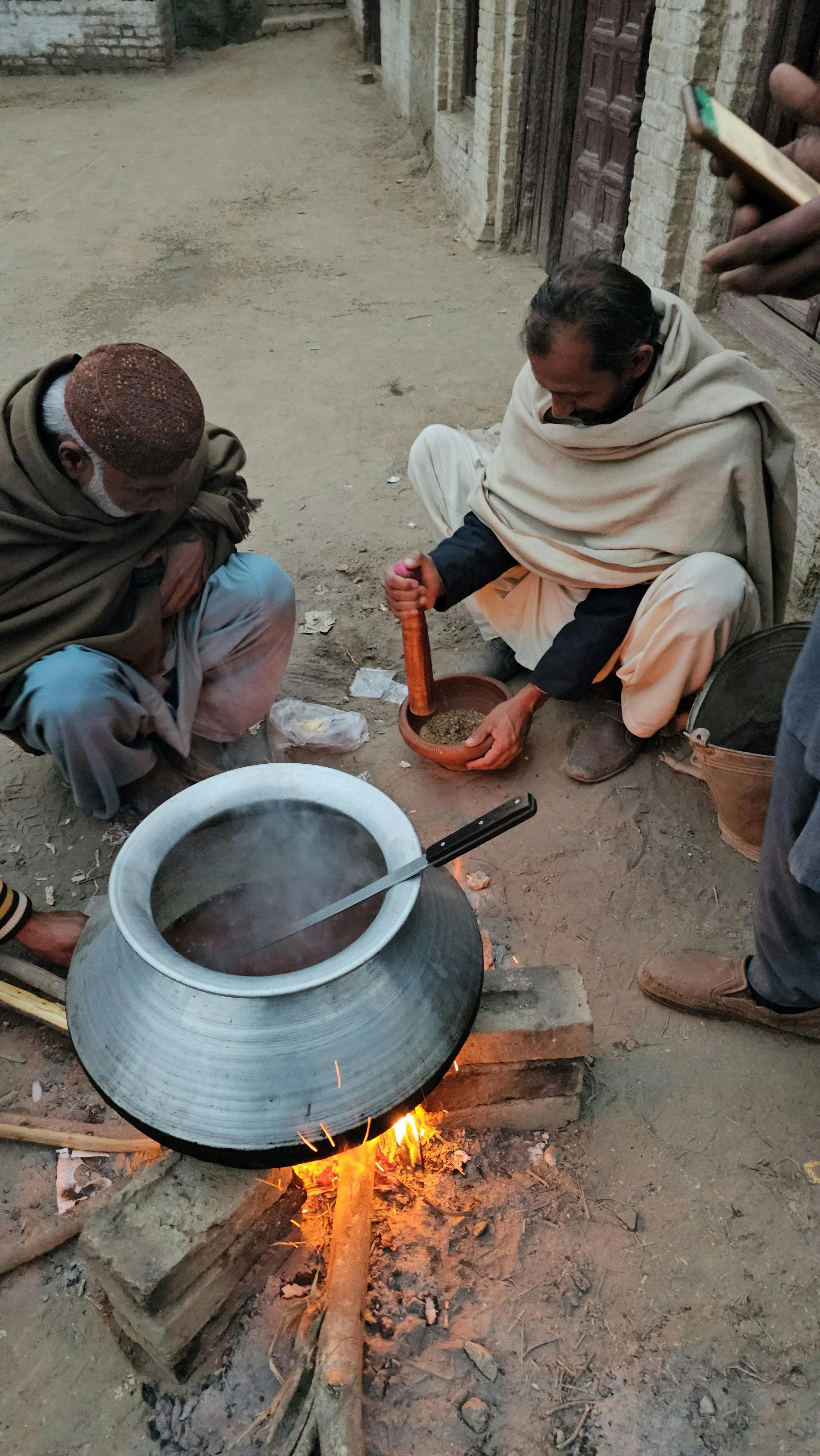 Two men prepare food outdoors with a large metal pot over open flames in a rustic village setting.
