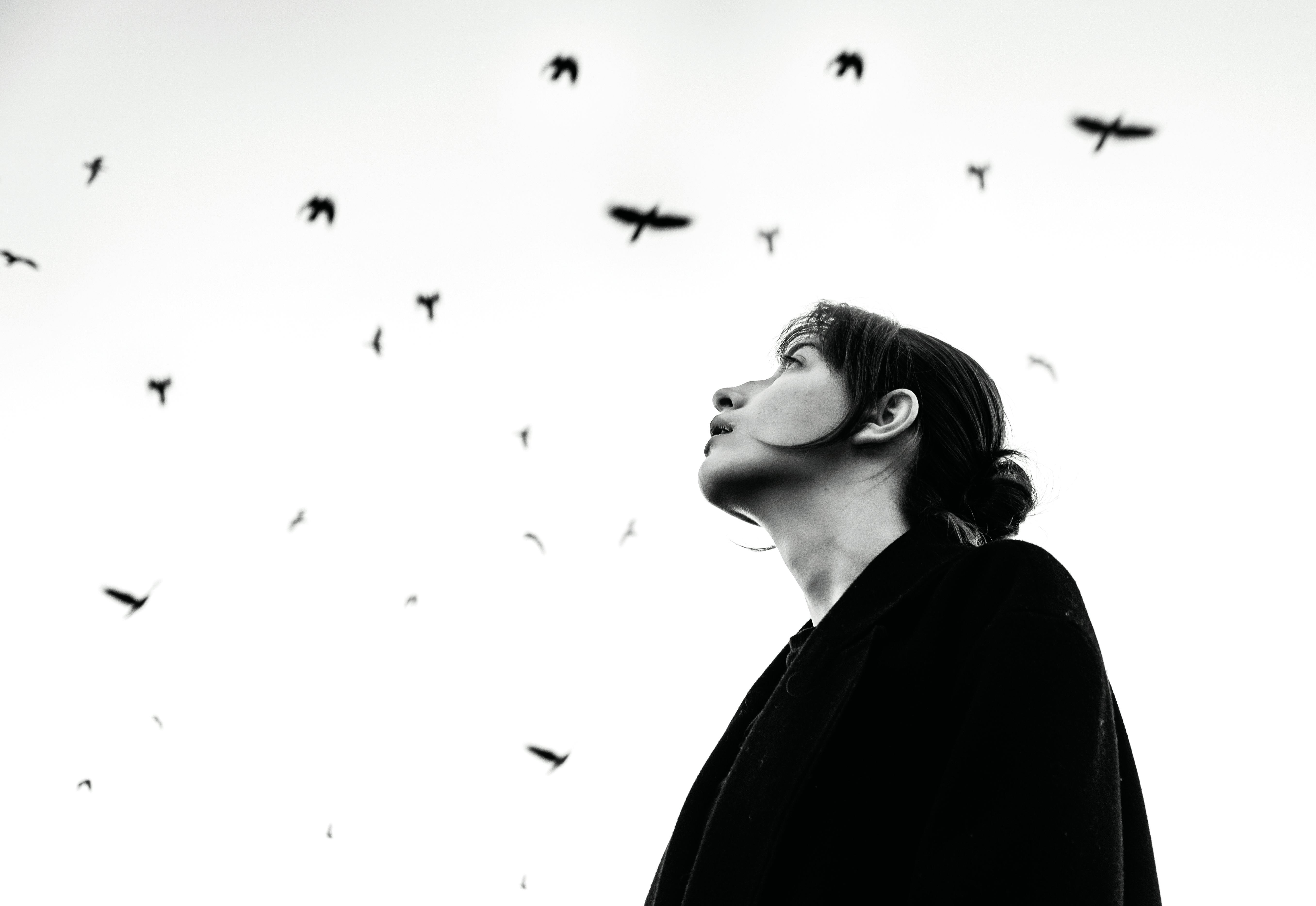 Artistic portrait of a woman looking up at birds in flight against a stark white sky.