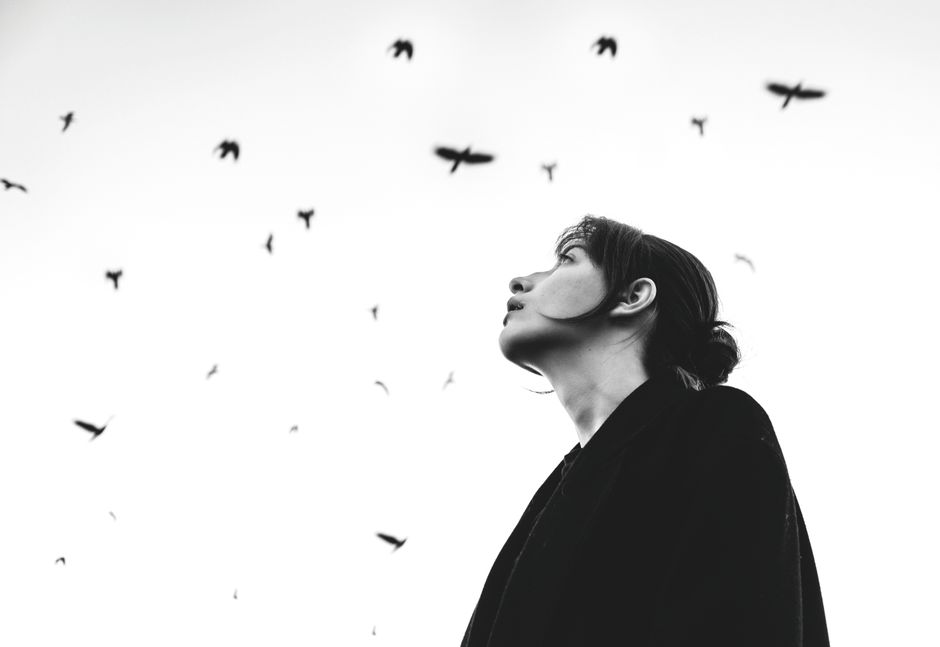 Artistic portrait of a woman looking up at birds in flight against a stark white sky.